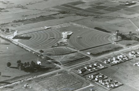 Black and white aerial photograph showing a square field used for a twin drive-in. The drive-ins each occupy a triangular half of the field. It is surrounded by other empty fields except for the bottom right where there are a series of houses. 